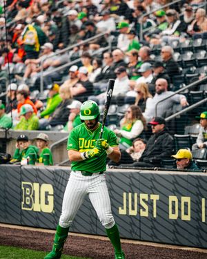 Oregon Ducks baseball player in green uniform batting at plate in Big Ten stadium with fans in background.