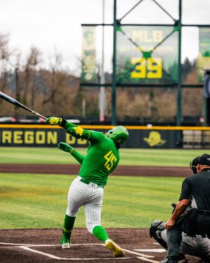 Oregon Ducks baseball player in green uniform swinging bat at PK Park, with MLB scoreboard showing number 39 in background.