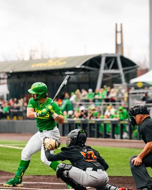 Oregon Ducks batter in green uniform swings at pitch during college baseball game with catcher and umpire behind home plate.