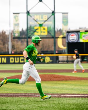 Oregon Ducks baseball player in green uniform runs bases at PK Park stadium with MLB scoreboard in background.