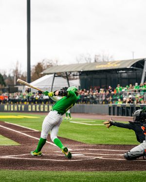 Oregon Ducks batter swings at pitch during college baseball game at PK Park, with catcher and crowd visible.
