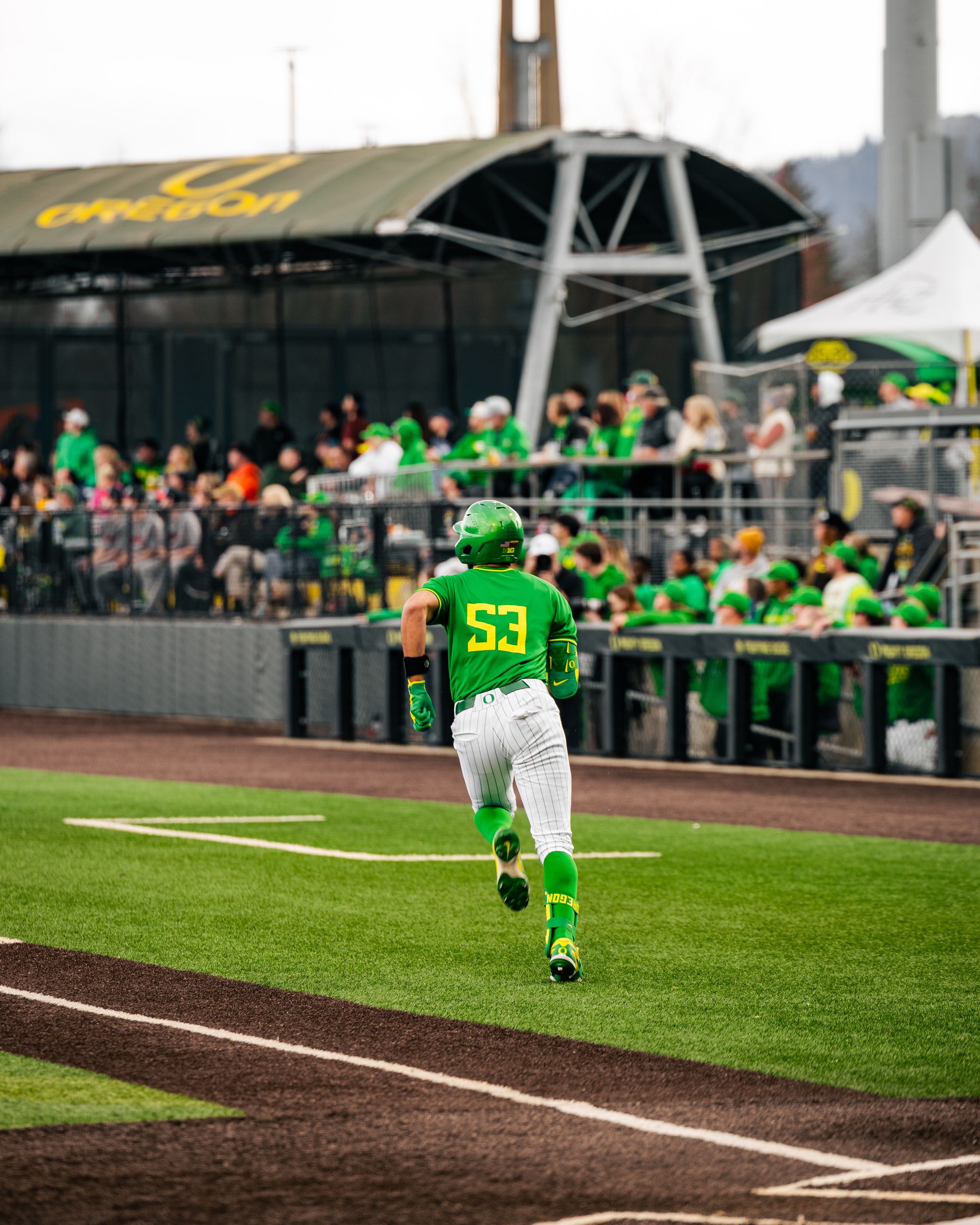 Oregon Ducks baseball player #53 in green uniform running bases at PK Park stadium with fans in background.