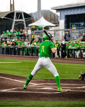 Oregon Ducks baseball player #11 in green uniform batting at home plate during a college game at a packed stadium.