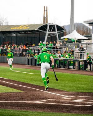 Oregon Ducks baseball player #11 in green uniform walks toward home plate carrying a bat during a college game.