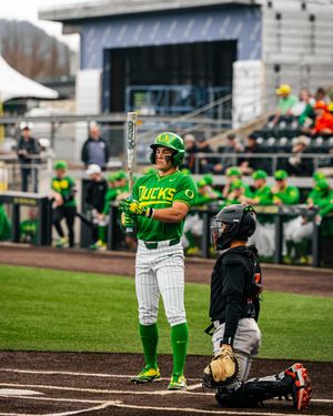 Oregon Ducks baseball player in green uniform holds bat at home plate during a college baseball game.