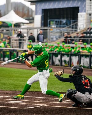 Oregon Ducks baseball player in green uniform swings bat during college game with catcher crouching behind home plate.