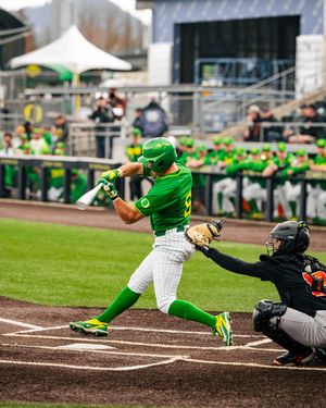 Oregon Ducks baseball player in green uniform swings bat during game while catcher crouches behind home plate.