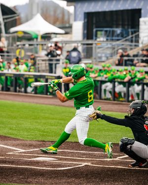 Oregon Ducks baseball player in green uniform swings bat during a college game, with catcher and dugout visible.