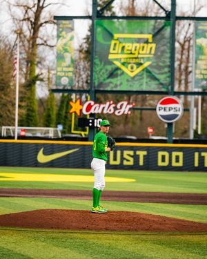 Oregon Ducks baseball pitcher stands on the mound at PK Park, wearing green uniform with Nike, Carl's Jr. and Pepsi ads visible.