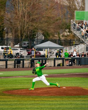 Oregon Ducks pitcher in green uniform delivers a pitch at a college baseball game with fans in the stands.
