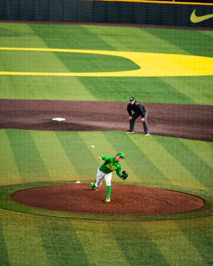 Oregon Ducks pitcher in green uniform delivers a pitch at PK Park baseball stadium with umpire watching.