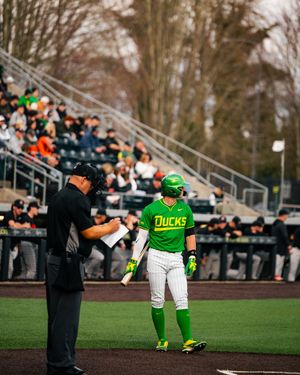 Oregon Ducks baseball player in green uniform talks with umpire during a game at a packed stadium.
