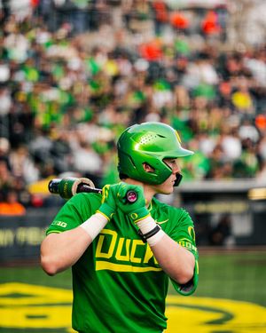 Oregon Ducks baseball player in green helmet and jersey holds bat ready at plate with crowd in background.