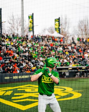 Oregon Ducks baseball player in green uniform batting at Bob Kilkenny Field with packed crowd in background.