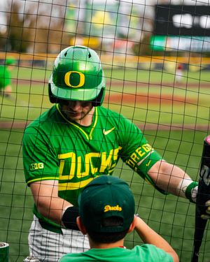 Oregon Ducks baseball player in green uniform and helmet signs autograph for young fan through batting cage net.