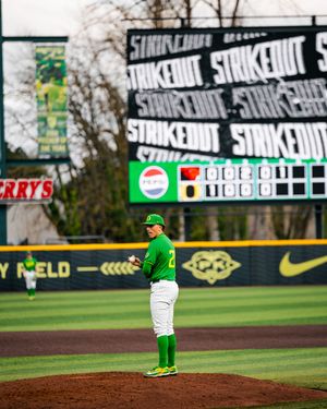 Oregon Ducks pitcher stands on mound in green uniform with strikeout scoreboard displayed at PK Park baseball stadium.