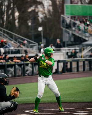 Oregon Ducks baseball player in green uniform batting at home plate during a college baseball game.