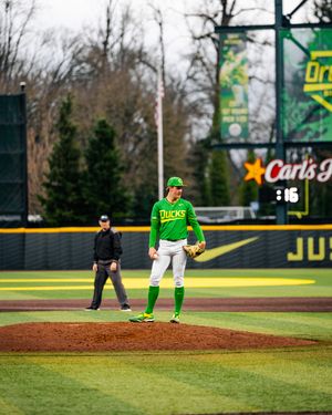 Oregon Ducks pitcher stands on the mound in green uniform at a college baseball game with umpire in background.