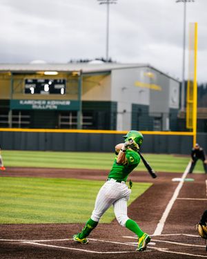 Oregon Ducks softball player in green uniform swings bat during a game at a modern baseball stadium.