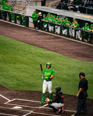Oregon Ducks baseball player stands at home plate holding a bat while teammates watch from the dugout.