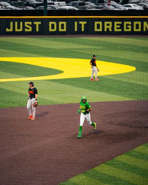 Oregon Ducks baseball players on field at PK Park with Just Do It Oregon sign visible in background.