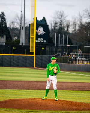 Oregon Ducks baseball pitcher stands on the mound in bright green uniform at PK Park, with Go Ducks sign visible.