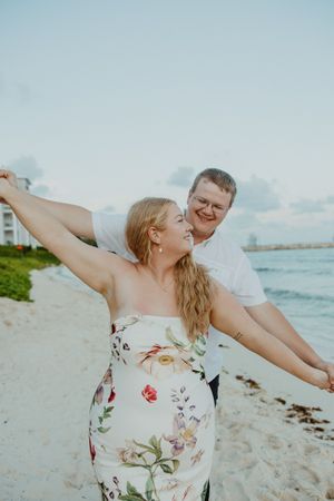 Couple dancing joyfully on a white sand beach, woman in floral dress with arms outstretched, man in white shirt behind her.