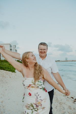 A couple dances joyfully on a sandy beach, woman in floral dress, man in white shirt, ocean waves in background.
