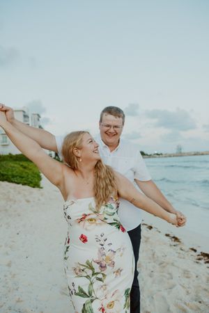 Couple dancing joyfully on a sandy beach, woman in floral dress with arms outstretched, man in white shirt, ocean backdrop.