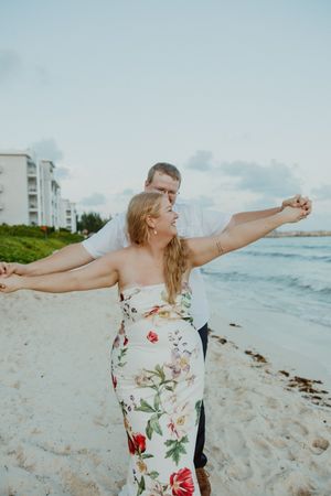 Couple dancing on a sandy beach, woman in floral dress with arms outstretched, ocean and resort in background.