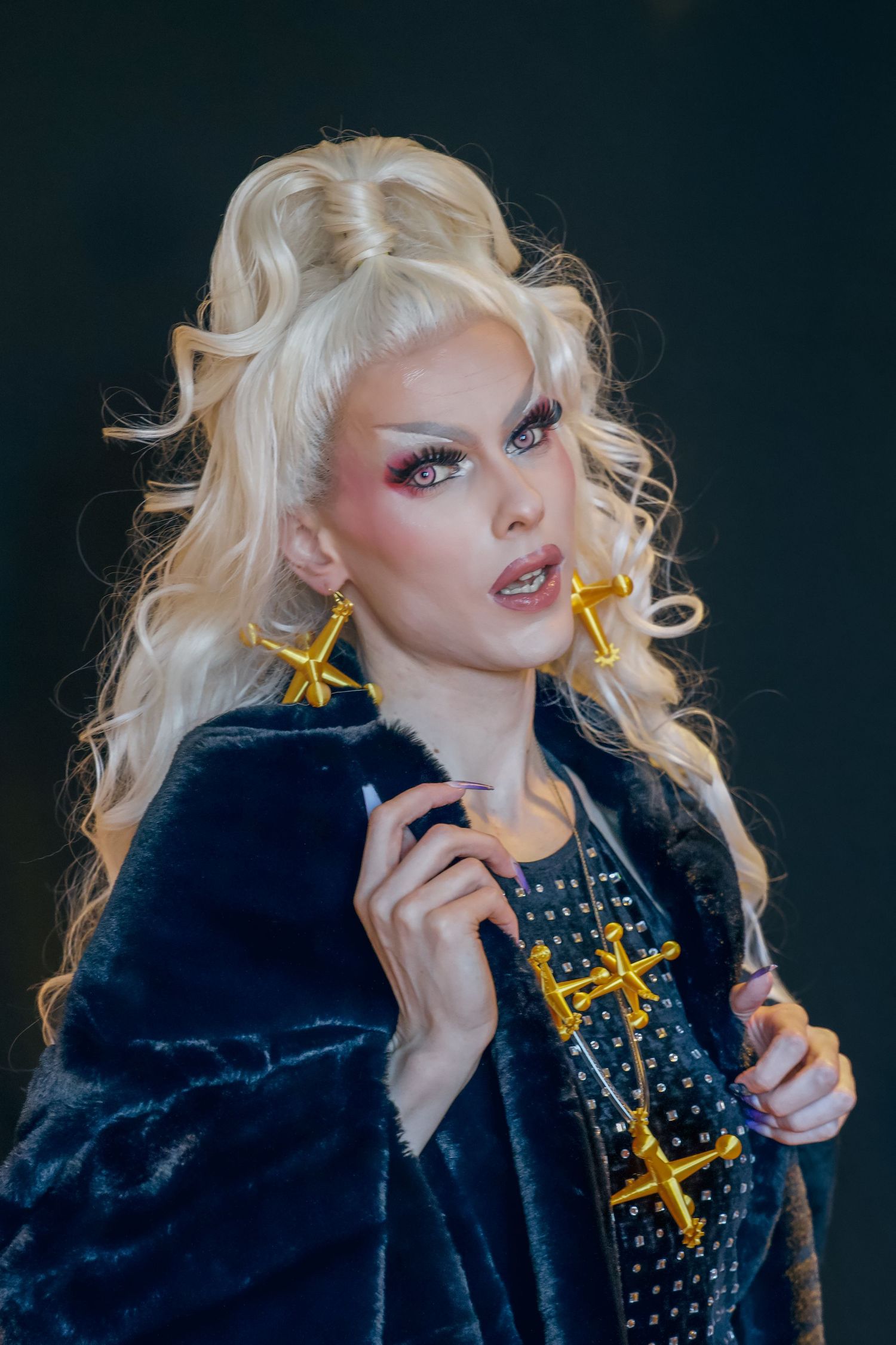 Drag performer with blonde curly hair, gold cross earrings, dramatic makeup, and black fur coat poses against dark background.
