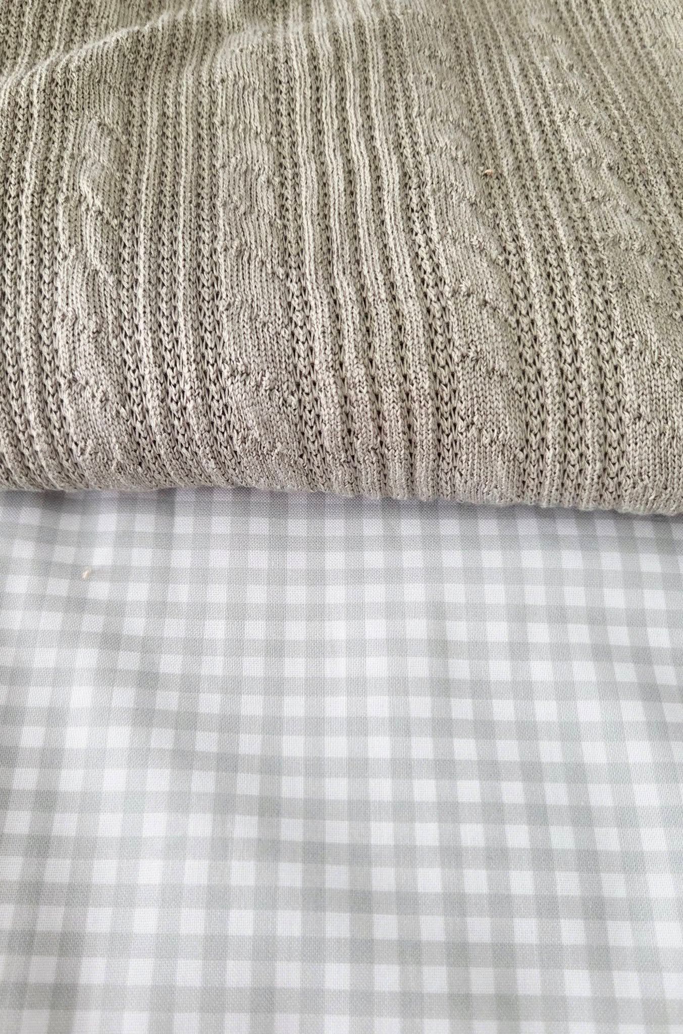 Beige cable knit blanket folded over gray and white gingham check fabric on a bed.
