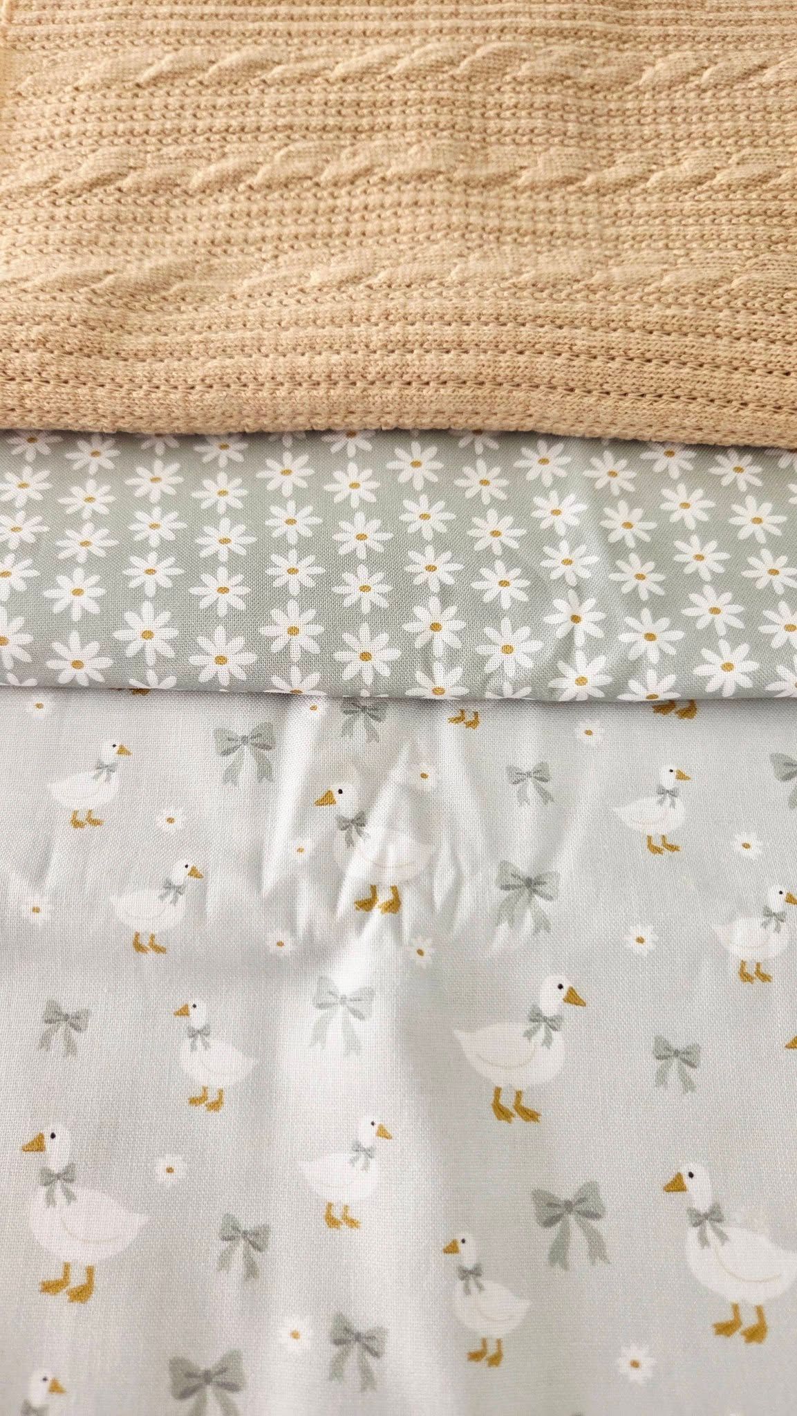 Three layered baby fabrics featuring a beige cable knit blanket, daisy print, and cute duck with bow pattern in soft grey tones.