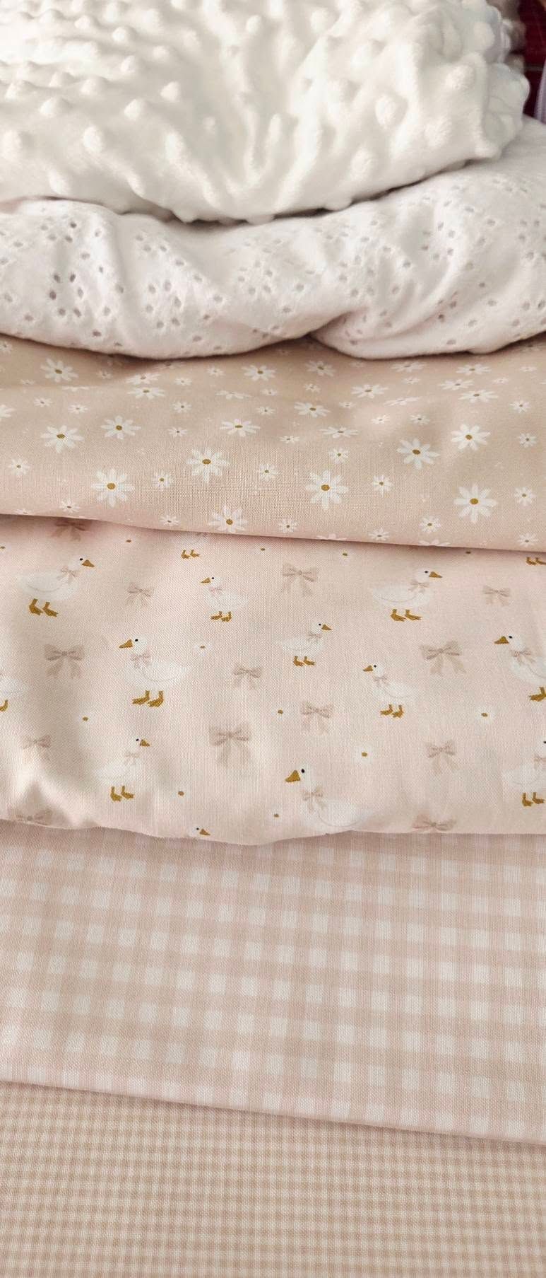 Stack of soft pink fabrics including eyelet lace, daisy print, duck and bow print, and gingham check patterns.