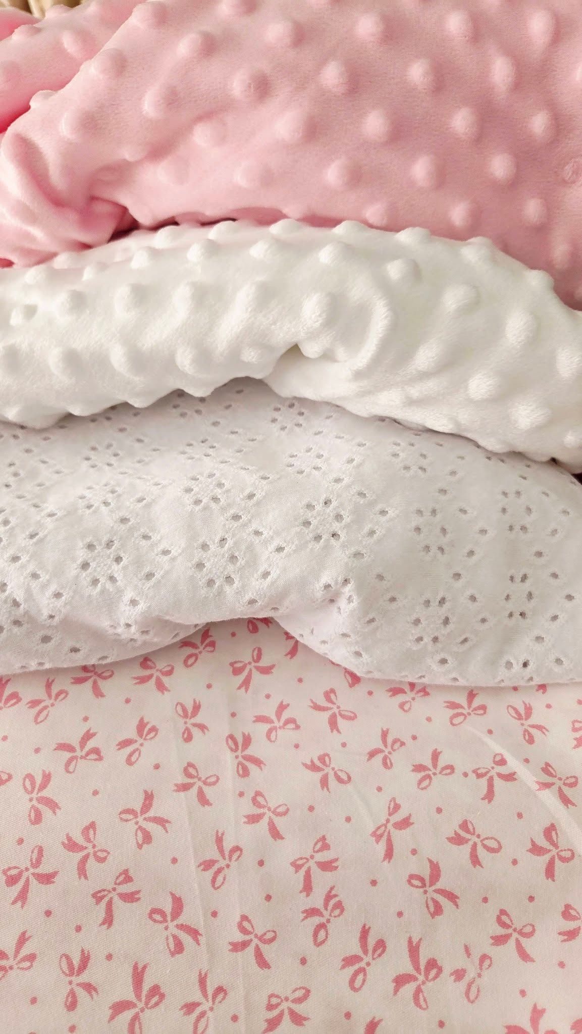 Layered pink and white minky dot, eyelet lace, and bow-print fabrics stacked together for a soft, feminine baby bedding display.