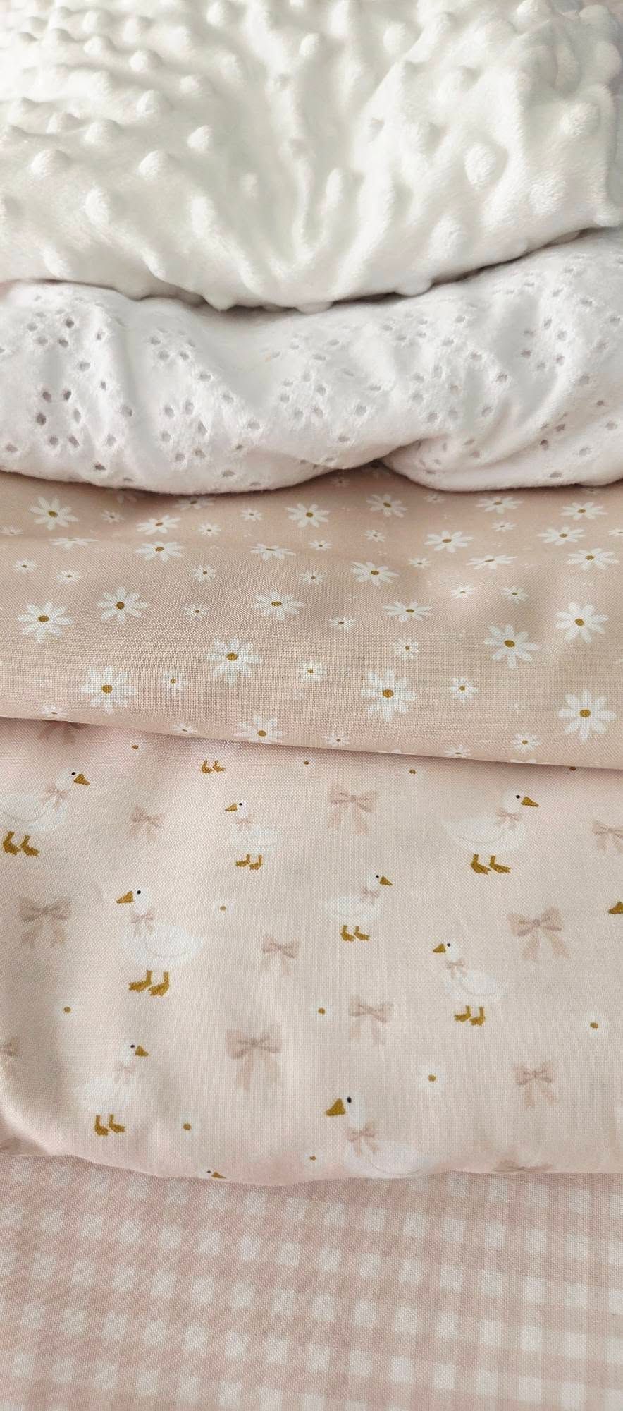 Stack of soft pink baby fabrics including minky dot, eyelet lace, daisy print, duck bow print, and pink gingham.