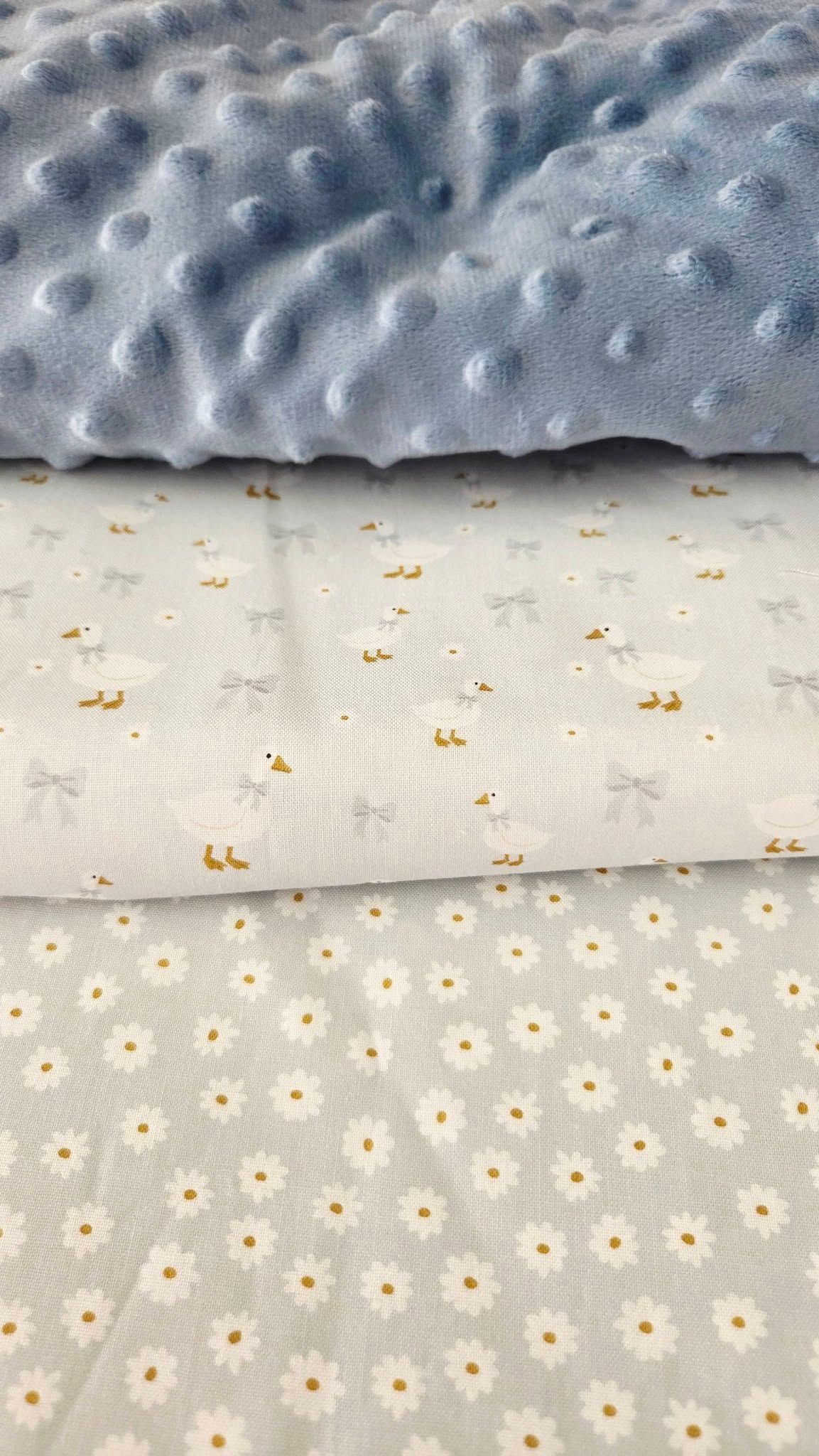 Three stacked baby fabrics: blue minky dot, white with ducks and bows, and white daisy print cotton.