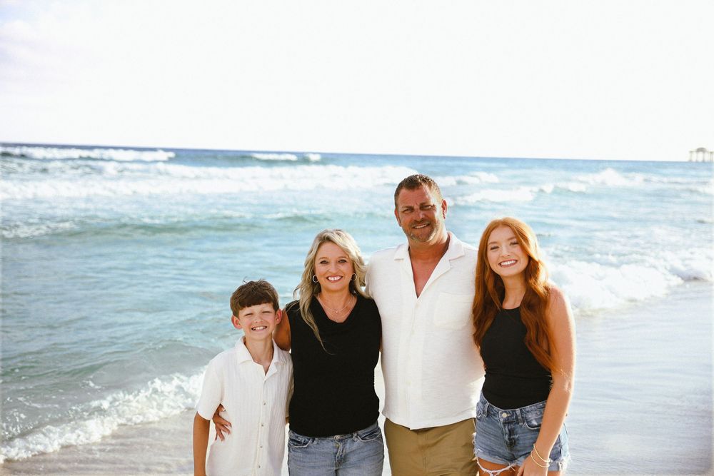 Family of four smiling together on a sunny beach with ocean waves in the background.