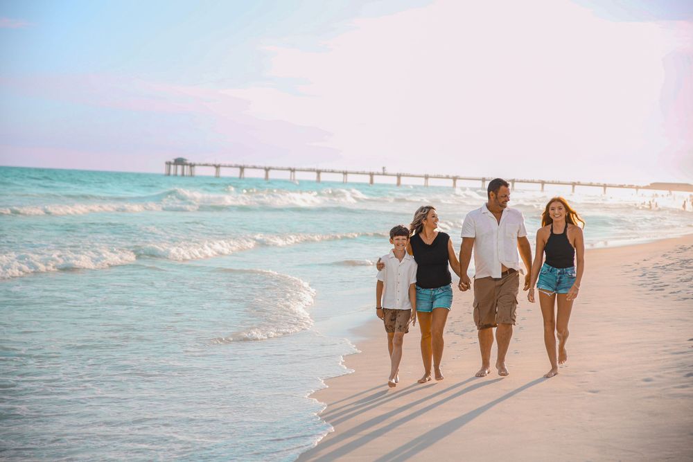 Family of four walking hand-in-hand on a sunny beach with turquoise waves and a pier in the background.