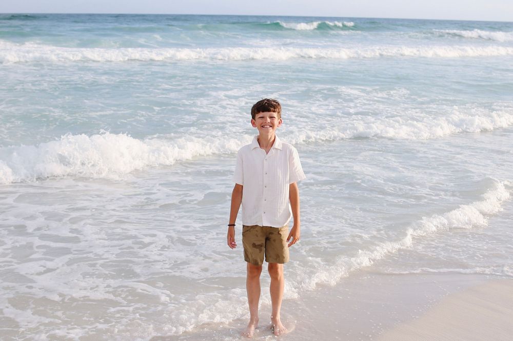 A smiling boy in a white shirt and khaki shorts stands barefoot on a sunny beach with ocean waves behind him.