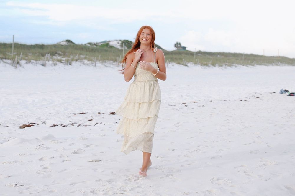 A young redhead woman in a flowing cream tiered dress walks barefoot on a white sandy beach.