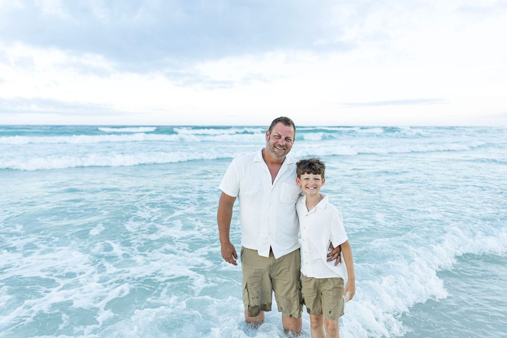 A man and young boy smile together on a sandy beach with ocean waves behind them on a cloudy day.