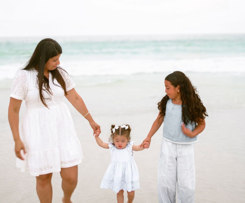A mother and two daughters hold hands and walk along a sandy beach, dressed in white and light blue outfits.