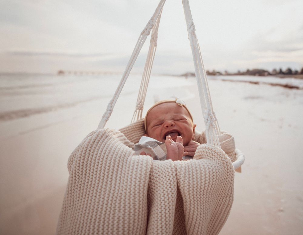 Newborn baby smiling in a macrame swing on a beach, wrapped in a cozy cream knit blanket.