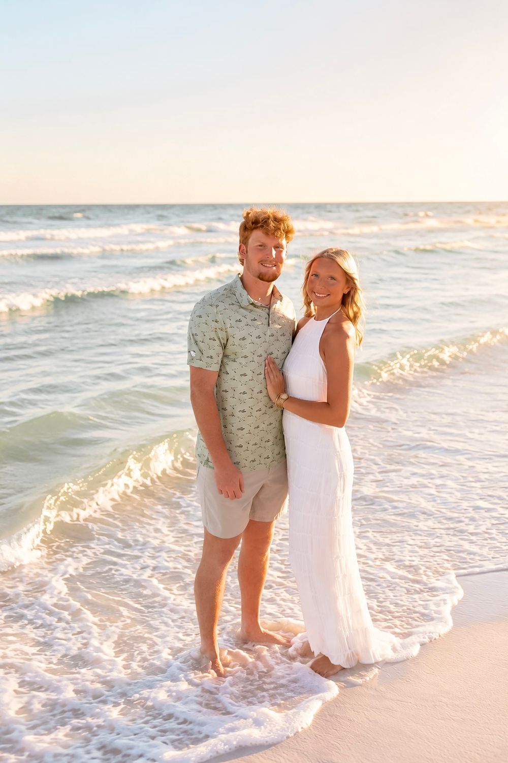 Couple smiling together on a sandy beach at sunset, man in green shirt and woman in white dress with ocean waves.