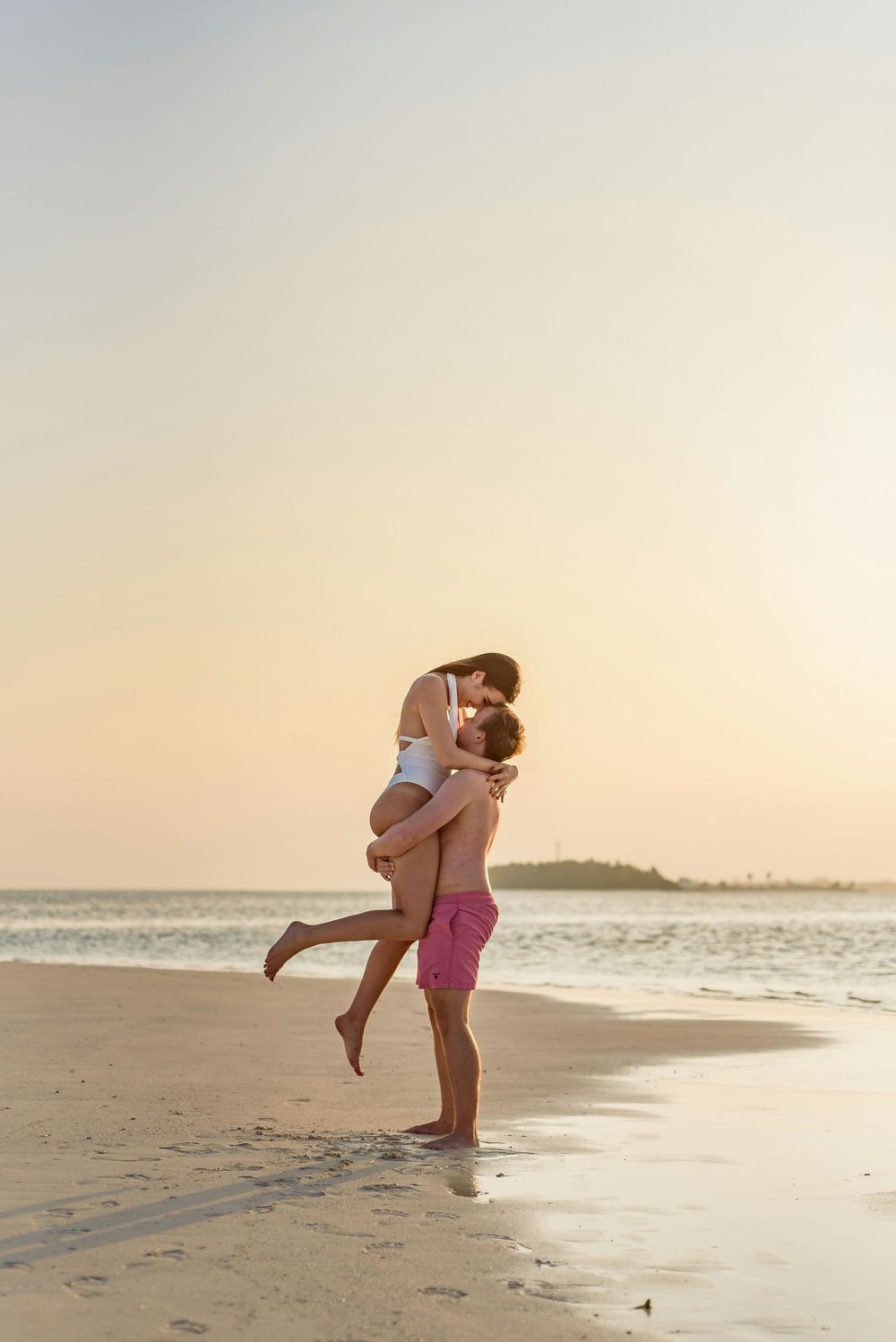 A man lifts and kisses a woman on a tropical beach during a warm golden sunset by the ocean.