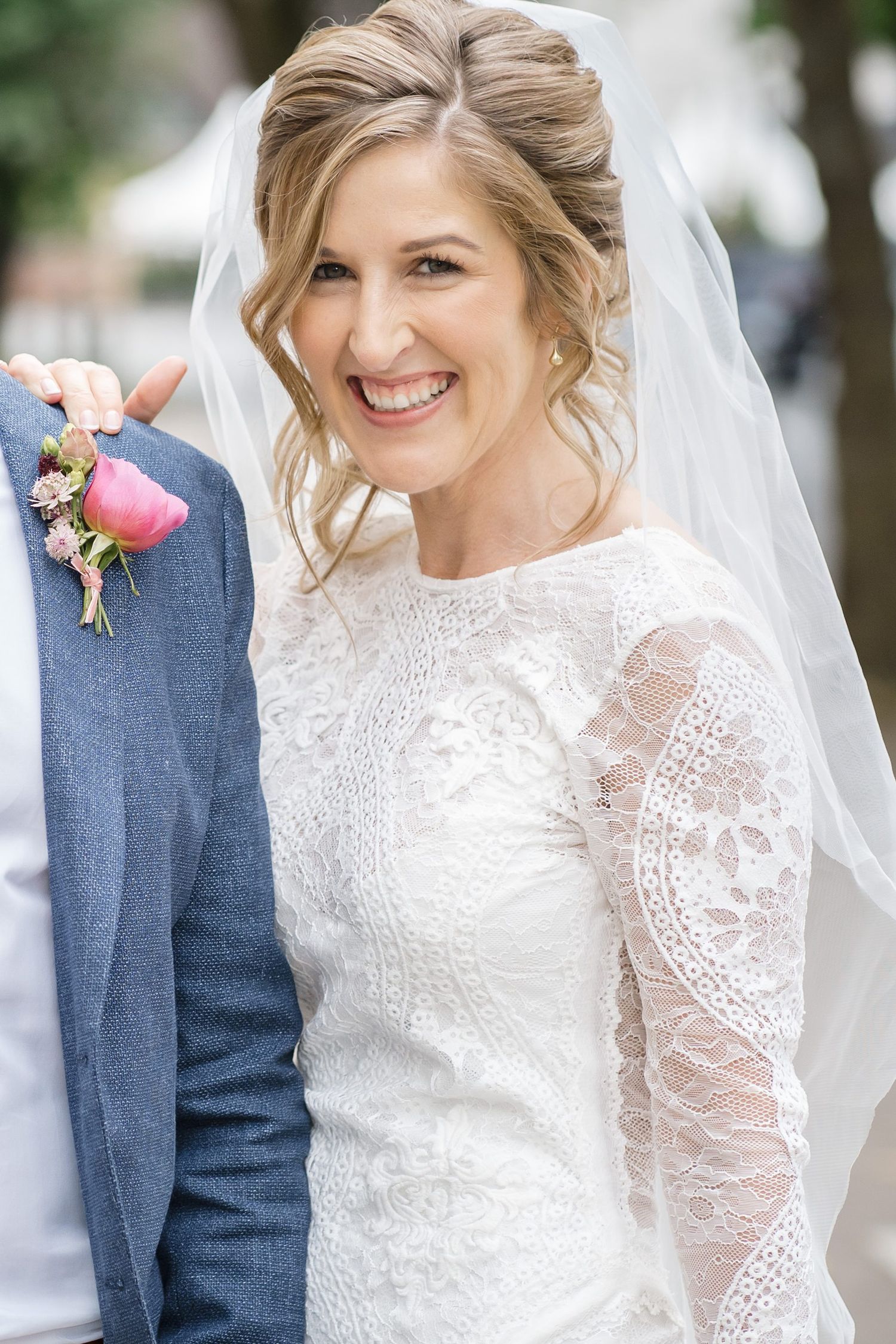 Smiling bride in lace long-sleeve wedding dress and veil, holding groom's arm with pink boutonniere outdoors Saraguay House