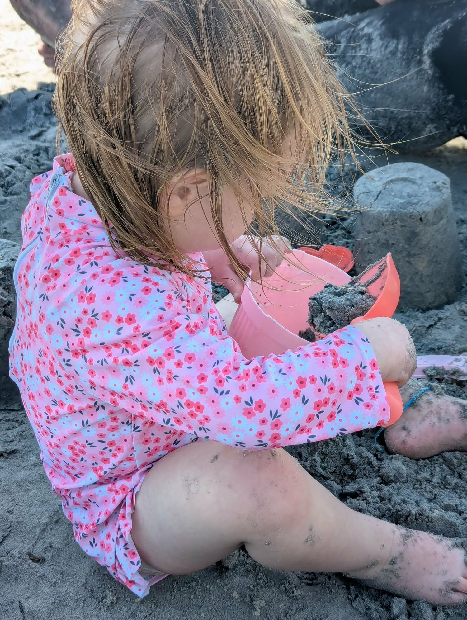 Young girl in floral swimsuit playing with wet sand and pink bucket at the beach.