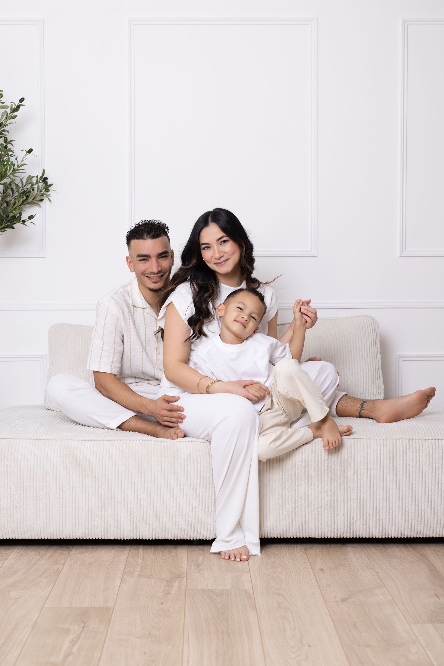 Happy family of three in white outfits sitting together on a cream sofa in a bright, elegant living room.