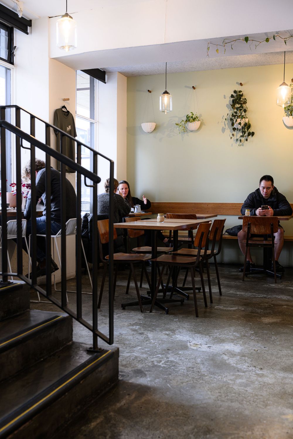 Modern cafe interior with wooden stairs, pendant lights, and concrete floors showing customers at tables.